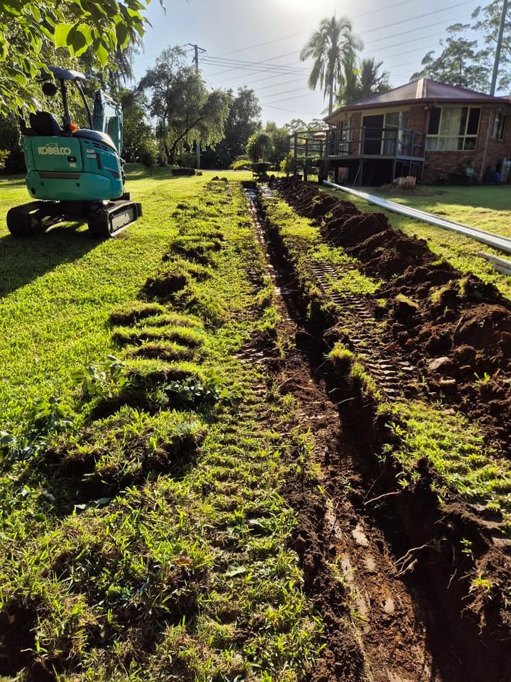Bonville stormwater trench at golden hour — Kobelco 3-tonne excavator on a green Coffs Coast lawn, freshly cut trench running across the yard.