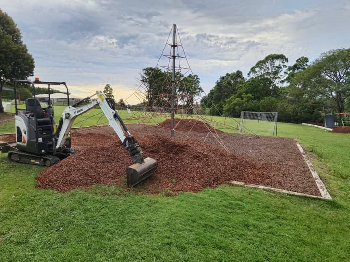 Mini-excavator mid-job at a school playground, mound of softfall bark around a climbing frame, late-afternoon Coffs Coast light.